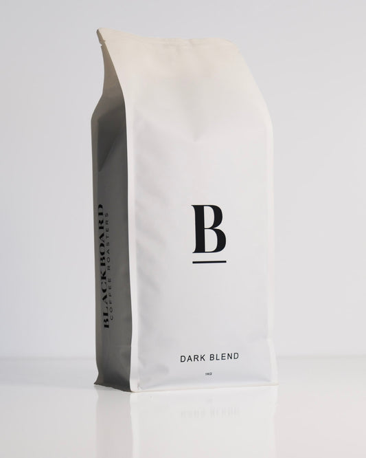 Roasted coffee beans. Blackboard Coffee Roasters, Blackboard Dark Espresso Blend. Image shows a white 1kg bag against a white background. The bag features the Blackboard logo centred in on the bag and the name of the blend centred at the bottom. The Roast Collective.