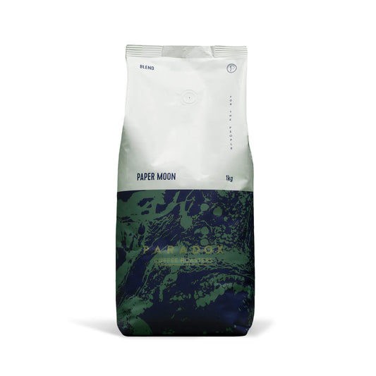 Roasted coffee beans from The Roast Collective. Paradox Coffee Roasters, Paper Moon Blend. Image shows a tri-tone 1kg bag of the coffee against a white background. The top half of the bag is white, with a navy blue and forest green pattern covering the bottom half of the bag. Paradox's logo is centred in the bottom half of the bag, with the lend name and weight at the bottom of the white section of the bag.