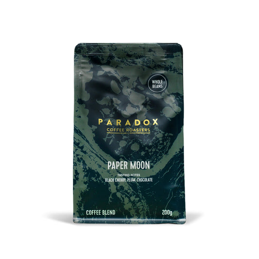 Roasted coffee beans from The Roast Collective. Paradox Coffee Roasters, Paper Moon Blend. Image shows a 200g bag of the coffee against a white background. The bag is two-tone in colour, with navy blue and forest green arranged in a organic pattern. Paradox's logo is centred in the top half of the bag, with the blend name, tasting notes and weight underneath.