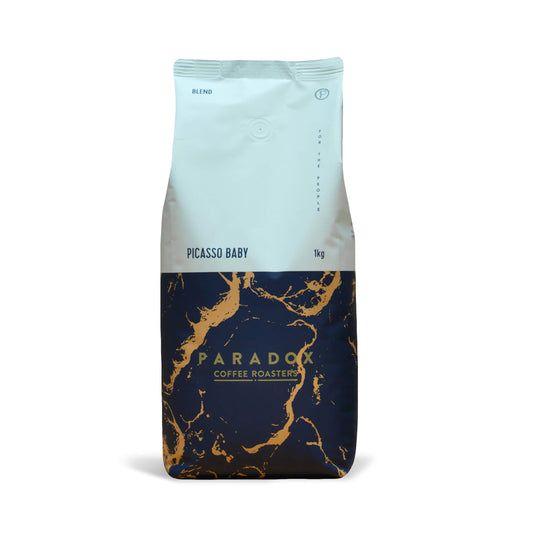 Roasted coffee beans from The Roast Collective. Paradox Coffee Roasters, Picasso Baby Blend. Image shows a tri-tone 1kg bag of the coffee against a white background. The top half of the bag is white, with a navy blue and gold coloured pattern covering the bottom half of the bag. Paradox's logo is centred in the bottom half of the bag, with the lend name and weight at the bottom of the white section of the bag.