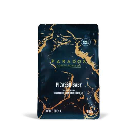 Roasted coffee beans from The Roast Collective. Paradox Coffee Roasters, Picasso Baby Blend. Image shows a 200g bag of the coffee against a white background. The bag is two-tone in colour, with navy blue and gold arranged in a geological print. Paradox's logo is centred in the top half of the bag, with the blend name, tasting notes and weight underneath.
