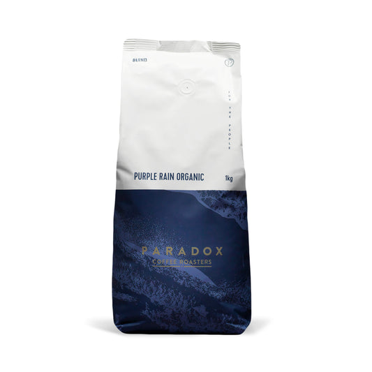 Roasted coffee beans from The Roast Collective. Paradox Coffee Roasters, Purple Rain Organic Blend. Image shows a tri-tone 1kg bag of the coffee against a white background. The top half of the bag is white, with a dark navy blue and deep purple pattern covering the bottom half of the bag. Paradox's logo is centred in the bottom half of the bag, with the lend name and weight at the bottom of the white section of the bag.