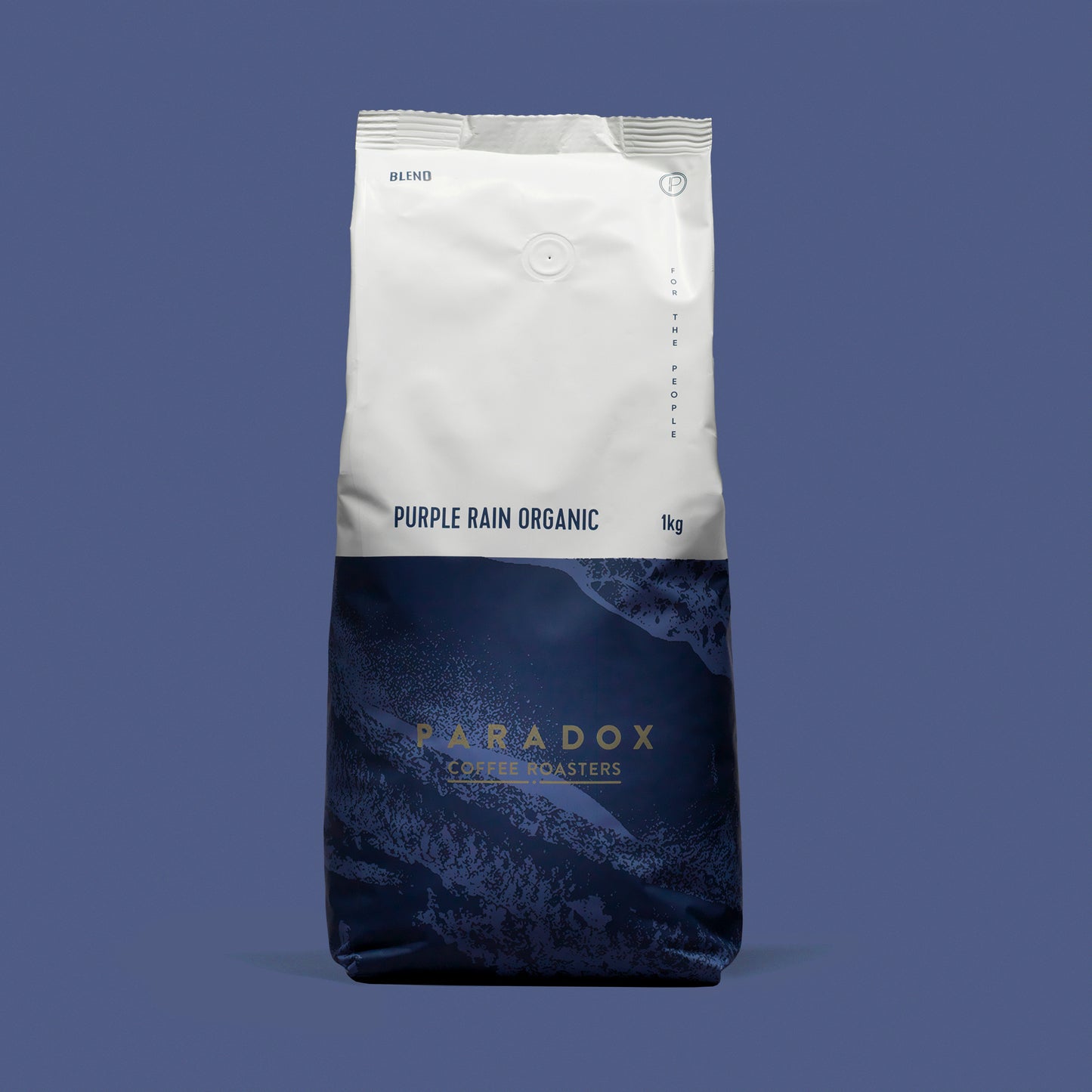 Roasted coffee beans from The Roast Collective. Paradox Coffee Roasters, Purple Rain Organic Blend. Image shows a tri-tone 1kg bag of the coffee against a grape purple coloured background. The top half of the bag is white, with a dark navy blue and deep purple pattern covering the bottom half of the bag. Paradox's logo is centred in the bottom half of the bag, with the lend name and weight at the bottom of the white section of the bag.