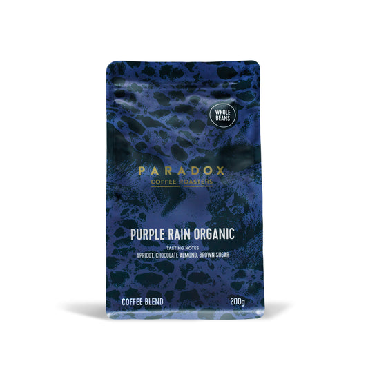 Roasted coffee beans from The Roast Collective. Paradox Coffee Roasters, Purple Rain Organic Blend. Image shows a 200g bag of the coffee against a white background. The bag is two-tone in colour, with deep purple and dark navy blue arranged in a reptile skin pattern. Paradox's logo is centred in the top half of the bag, with the blend name, tasting notes and weight underneath.