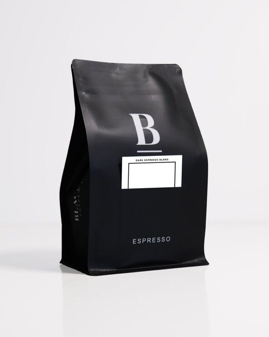 Roasted coffee beans. Blackboard Coffee Roasters, Blackboard Dark Espresso Blend. Image shows a black 250g bag against a white background. The bag features the Blackboard logo and a pouch on the front with a product information card poking out. The Roast Collective.