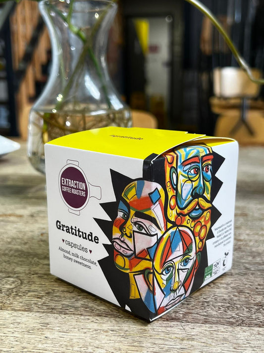 Extraction Coffee Roasters. Gratitude Capsules, for Nespresso compatible pod machines. The image shows a 10 pack box of capsules on a wooden table with a glass vase blurred in the background. The Roast Collective.