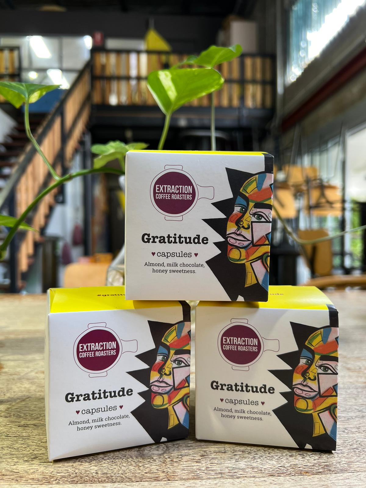 Extraction Coffee Roasters. Gratitude Capsules, for Nespresso compatible pod machines. The image shows three 10 pack boxes of capsules stacked like a pyramid on a wooden table with a plant and the roastery blurred in the background. The Roast Collective.