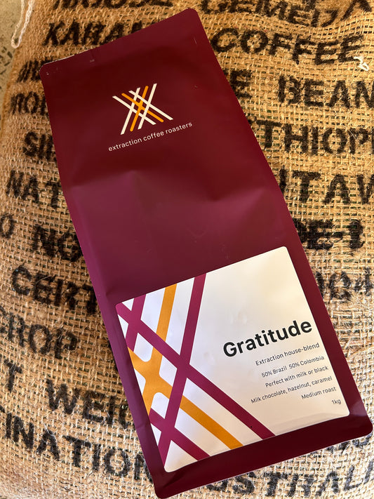 Roasted coffee beans. Extraction Coffee Roasters, Gratitude Blend. Image shows a 1kg bag of gratitude blend laying on top of a burlap coffee sack. The bag is maroon in colour. The Roast Collective