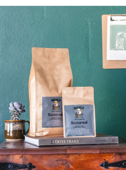 Roasted coffee beans from The Roast Collective. That's Whyld Coffee Roasters, Nocturnal Organic Blend. Image shows a 250g and a 1kg bag of Nocturnal blend next to each other, staged on a book titled "Coffee Trails", next to a succulent grown in a coffee cup. These are on the top of a wooden chest in front of a ocean green coloured background. The bags each have a grey blue label centred on the lower half of the bag with the roaster's logo, blend name, and blend details on it.