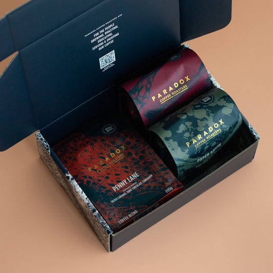 Roasted coffee beans from The Roast Collective. Paradox Coffee Roasters, Trio Blend Box. Image shows a blue and white Paradox-branded box containing 
 a 200g bag each of Paradox's Penny Lane, Paper Moon, and Poker Face blends. The box is positioned at a right leaning diagonal angle in the image against a beige coloured background.