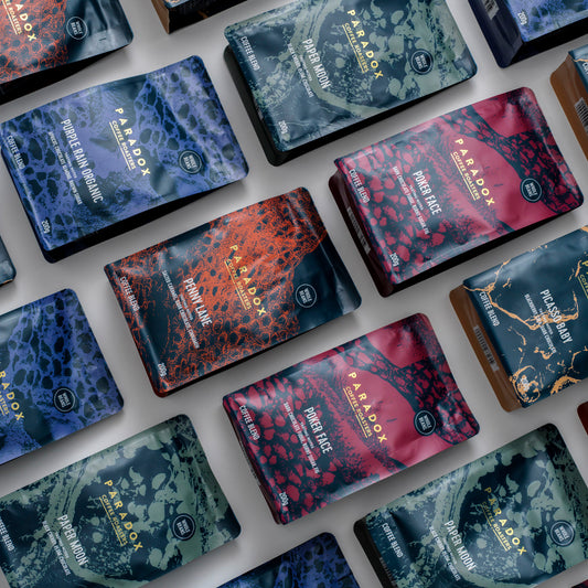Roasted coffee beans from The Roast Collective. The image shows 200g bags of each of Paradox's espresso blends, organised in a brick-like pattern diagonally across the images. The bags are interspersed by approximately 2 centimetres and are laid against a grey-white background. For more details about the colour and design of each bag, please see the individual bag image descriptions in this product.