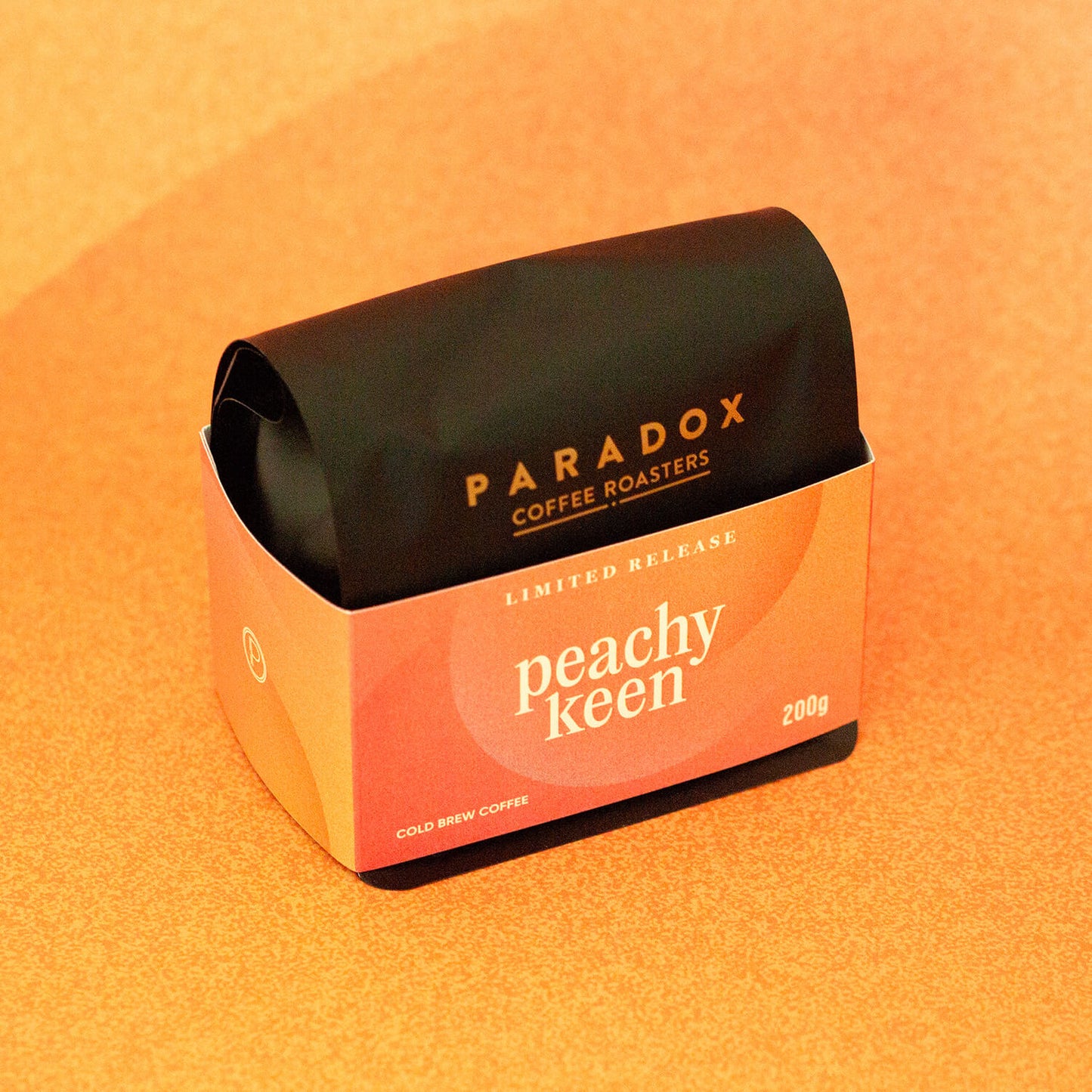 Roasted coffee beans Paradox Coffee Roasters Limited Edition: Peachy Keen 200g bag Cold Brew Coffee. Peach and orange coloured background, taken from an angle. The Roast Collective.