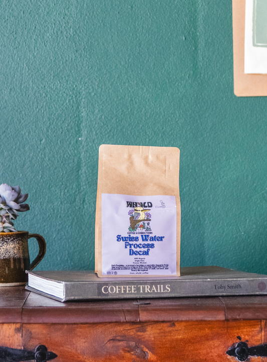 Roasted coffee beans from The Roast Collective. That's Whyld Coffee Roasters, Café Feminino Organic Decaf. Image shows a brown 250g bag of Café Feminino staged on a book titled "Coffee Trails", next to a succulent grown in a coffee cup. These are on the top of a wooden chest in front of a ocean green coloured background. The bag has a lavender coloured label centred on the lower half of the bag with the roaster's logo, blend name, and blend details on it.