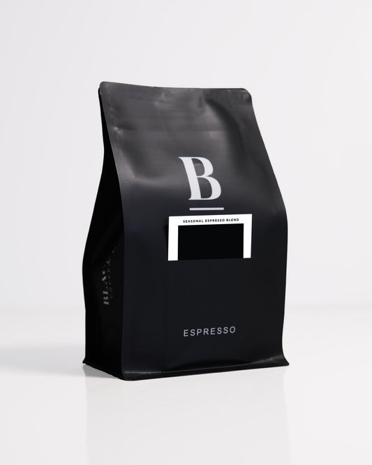 Roasted coffee beans. Blackboard Coffee Roasters, Blackboard Seasonal Espresso Blend. Image shows a black 250g bag against a white background. The bag features the Blackboard logo and a pouch on the front with a product information card poking out. The Roast Collective.
