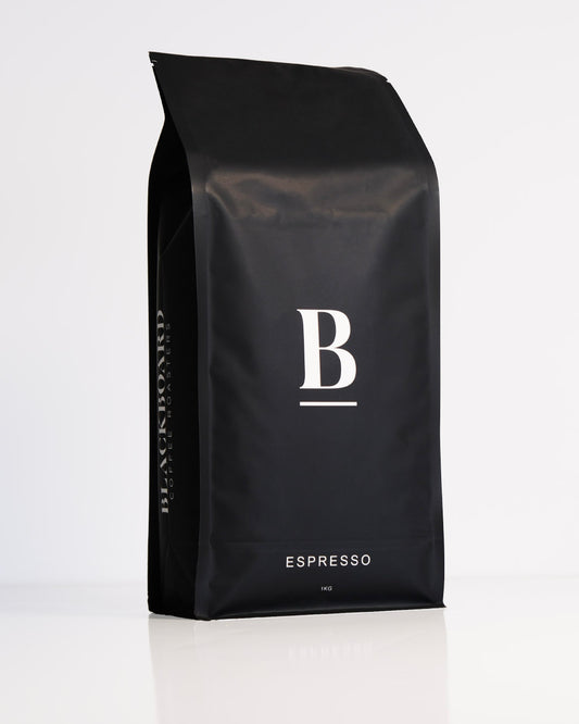 Roasted coffee beans. Blackboard Coffee Roasters, Blackboard Dark Espresso Blend. Image shows a black 1kg bag against a white background. The bag features the Blackboard logo centred in the middle of the bag and the blend name centred at the bottom. The Roast Collective.