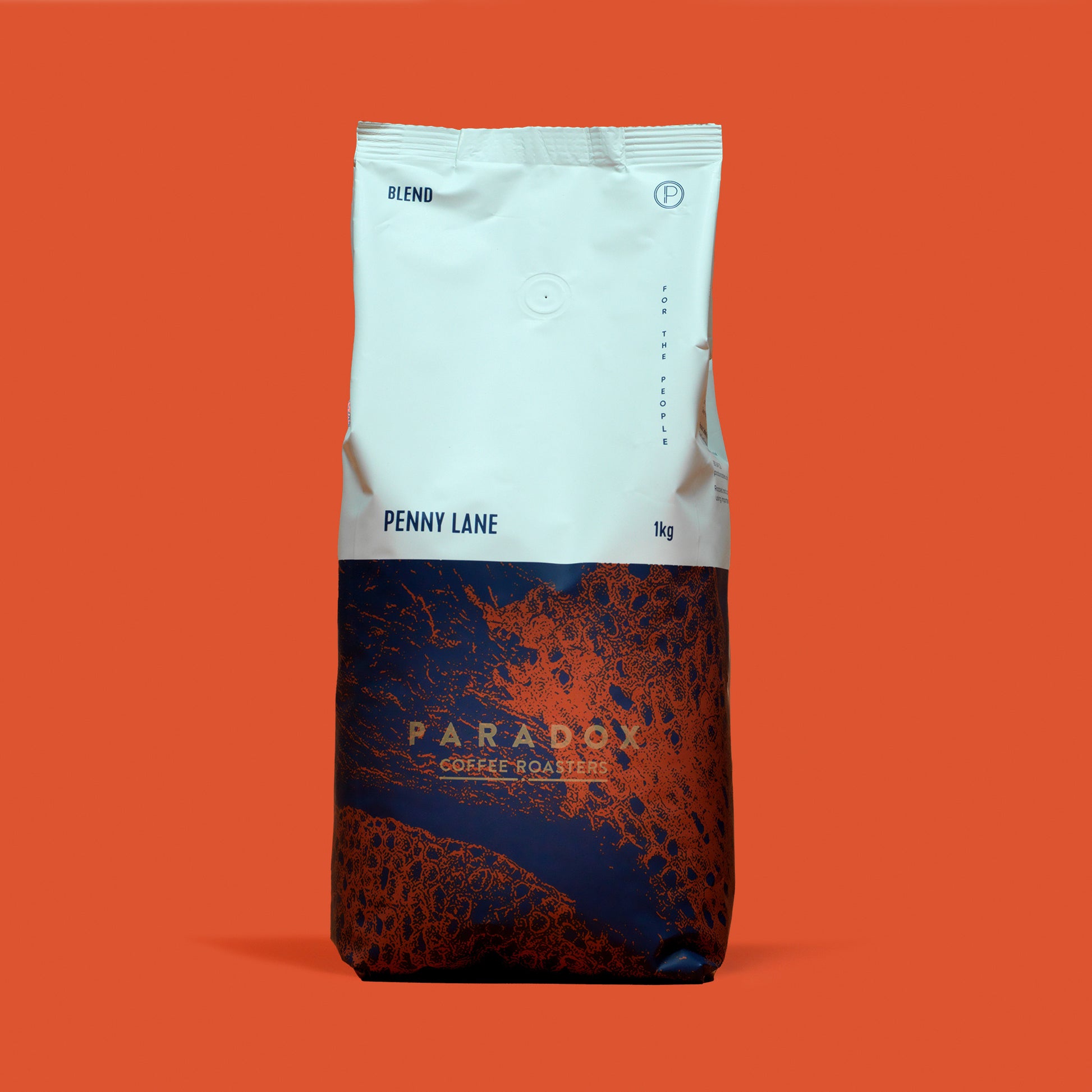 Roasted coffee beans from The Roast Collective. Paradox Coffee Roasters, Penny Lane Blend. Image shows a tri-tone 1kg bag of the coffee against a burnt orange coloured background. The top half of the bag is white, with a navy blue and rust red pattern covering the bottom half of the bag. Paradox's logo is centred in the bottom half of the bag, with the lend name and weight at the bottom of the white section of the bag. 