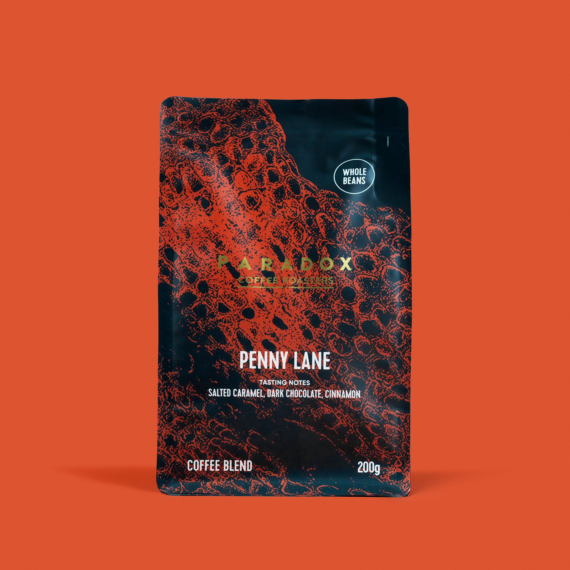 Roasted coffee beans from The Roast Collective. Paradox Coffee Roasters, Penny Lane Blend. Image shows a 200g bag of the coffee against a burnt orange coloured background. The bag is two-tone in colour, with navy blue and rust red arranged in a crocodile skin pattern. Paradox's logo is centred in the top half of the bag, with the blend name, tasting notes and weight underneath. 