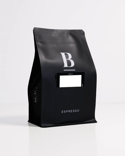 Roasted coffee beans. Blackboard Coffee Roasters, Blackboard Single Origin Decaf. Image shows a black 250g bag against a white background. The bag features the Blackboard logo and a pouch on the front with a product information card poking out. The Roast Collective.