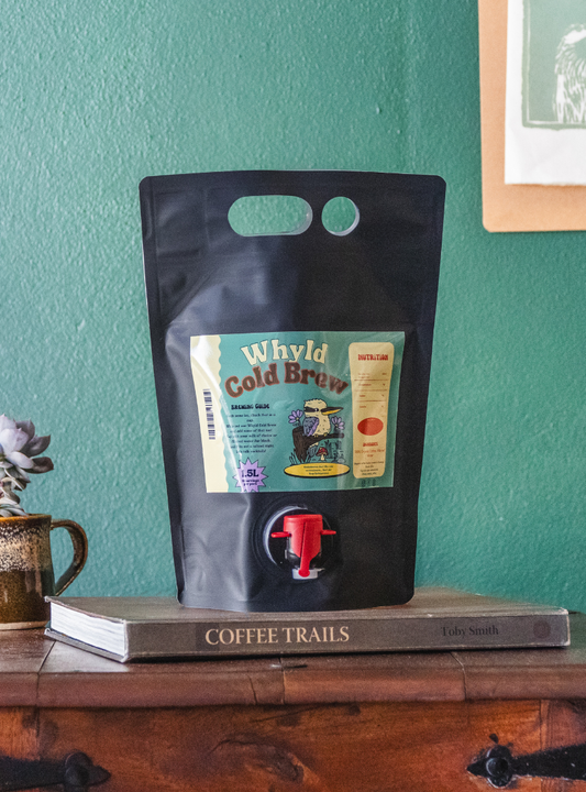 Ready to drink cold brew coffee from The Roast Collective. That's Whyld Coffee Roasters, Organic Cold Brew Goonie. Image shows a black 1.5L "goon bag" of  cold brew coffee staged on a book titled "Coffee Trails", next to a succulent grown in a coffee cup. These are on the top of a wooden chest in front of a ocean green coloured background. The bag has an ocean green coloured label centred in the middle of the bag with the product name and details details on it.