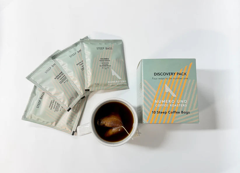 Coffee by The Roast Collective. Image shows a box of Numero Uno Coffee Roaster's Discovery Pack Steep Bags, along with four individual steep bag packets, arranged around a top-down view of a cup with one of the steep bags brewing inside. 
