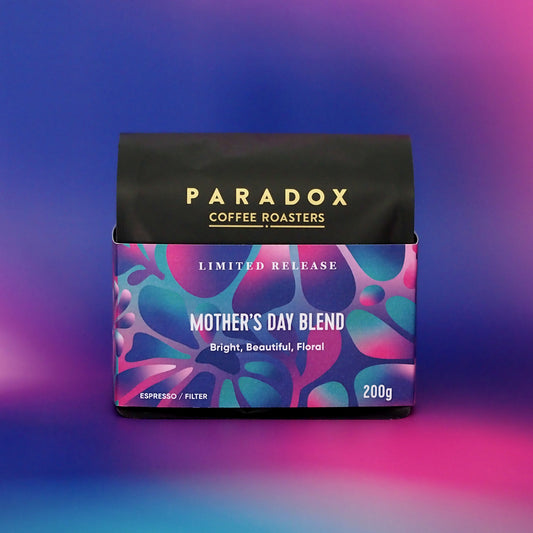 Roasted coffee beans. Paradox Coffee Roasters - Limited Edition Mother's Day Blend. Omni-roast: suitable for espresso and filter. Image shows a 200g bag facing front on against a pink, purple and blue pastel gradient patterned background. The Roast Collective.