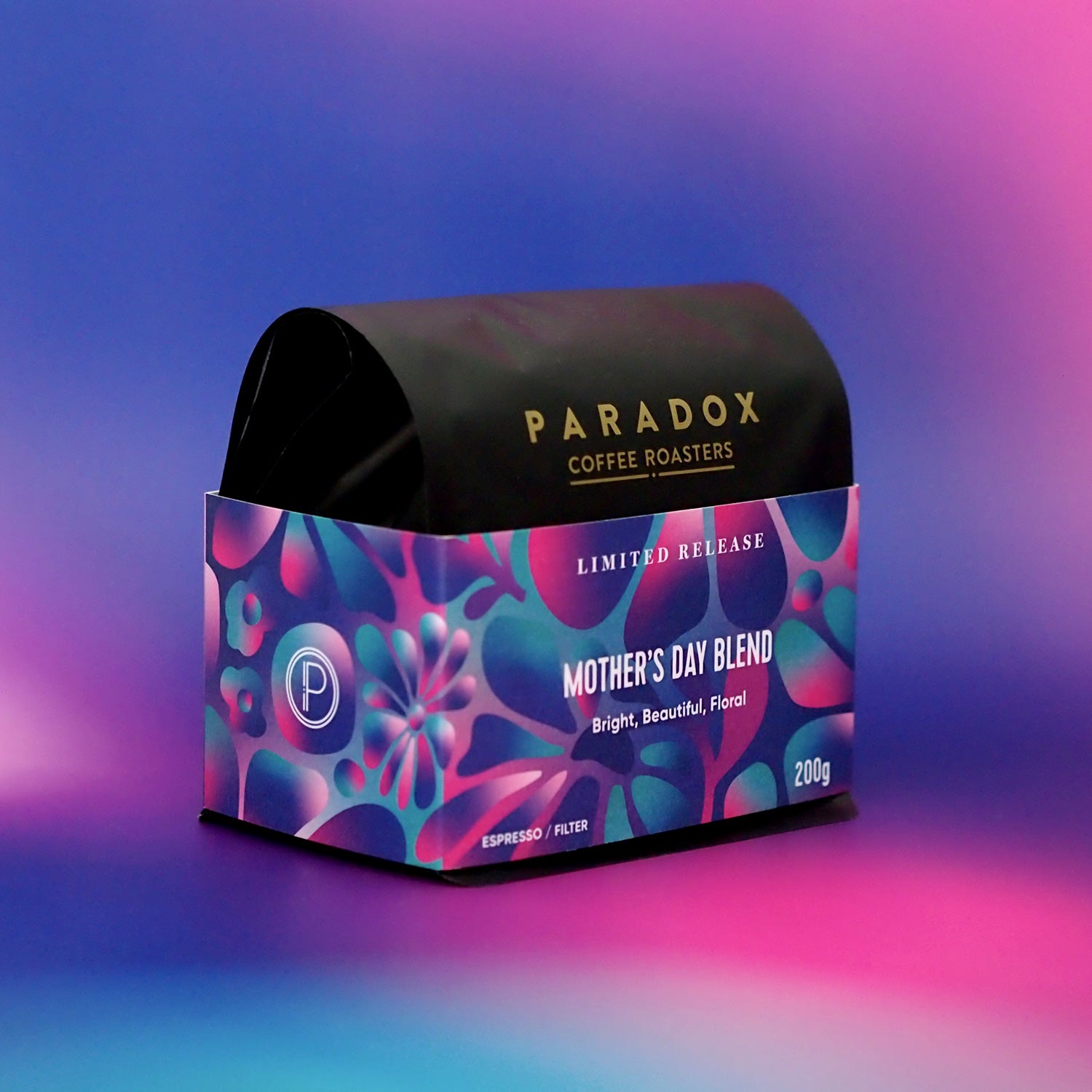 Roasted coffee beans. Paradox Coffee Roasters - Limited Edition Mother's Day Blend. Omni-roast: suitable for espresso and filter. Image shows a 200g bag facing at a partial right-sided side view against a pink, purple and blue pastel gradient patterned background. The Roast Collective.