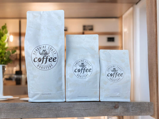Roasted coffee beans from The Roast Collective. Sunshine Coast Coffee Roastery, Sunset Blend. Image shows a 1kg, 500g, and 250g bag of Sunset blend stacked next to each other on a wooden table with the roastery store in the  background. The bags have the roaster's logo centred on the front of the bag.