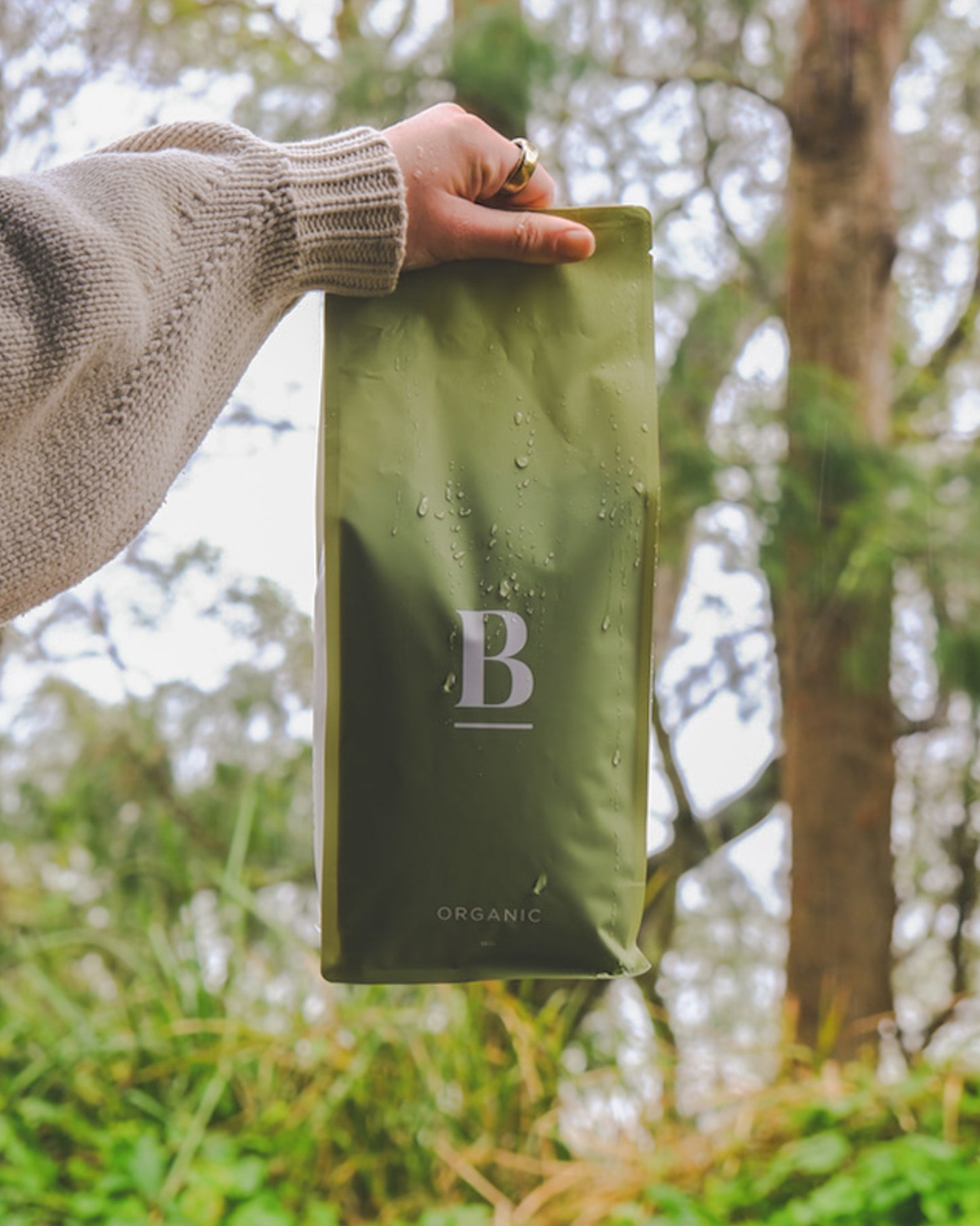 Roasted coffee beans. Blackboard Coffee Roasters, Blackboard Organic Espresso Blend. Image shows a green and white coloured 1kg bag being held at the top by a woman's hand against a blurred background of the Australian bush. Only the woman's hand and forearm are in frame. The bag has a few drops of water on the front, as if it's been hit by heavy raindrops and features the Blackboard logo centred in the middle of the bag and the blend name centred at the bottom. The Roast Collective. 