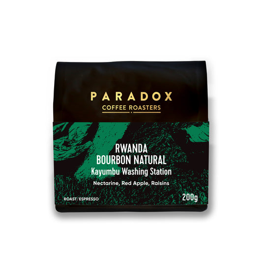 Roasted coffee beans from The Roast Collective. Paradox Coffee Roasters, Rwanda Bourbon Natural from Kayumbu Station. Image shows a black 200g bag of the coffee against a white background. The bag has Paradox's logo centred at the top of the bag in gold coloured print and a forest green coloured label centred on the lower half of the bag with the product name, tasting notes, roast profile and weight on it.