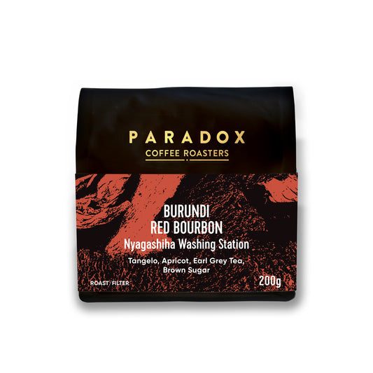 Roasted coffee beans from The Roast Collective. Paradox Coffee Roasters, Burundi, Red Bourbon by Nyagashiha Station. Image shows a black 200g bag of the coffee against a white background. The bag has Paradox's logo centred at the top of the bag in gold coloured print and a rust red coloured label centred on the lower half of the bag with the product name, tasting notes, roast profile and weight on it.
