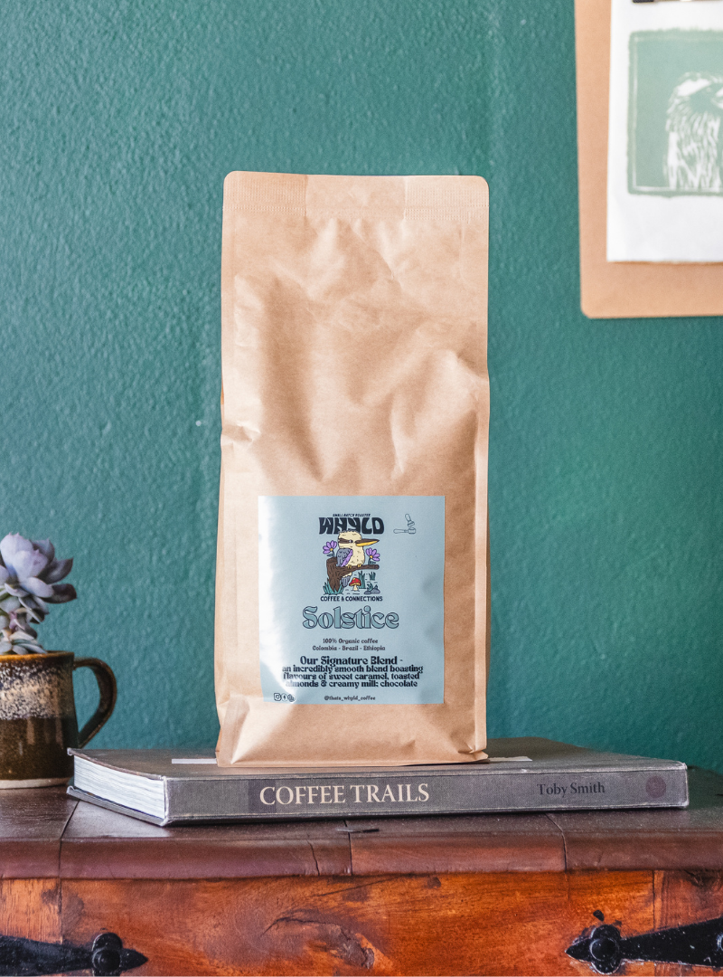 Roasted coffee beans from The Roast Collective. That's Whyld Coffee Roasters, Solstice Organic Blend. Image shows a brown 1kg bag of Solstice blend staged on a book titled "Coffee Trails", next to a succulent grown in a coffee cup. These are on the top of a wooden chest in front of a ocean green coloured background. The bag has a duck egg blue coloured label centred on the lower half of the bag with the roaster's logo, blend name, and blend details on it. 