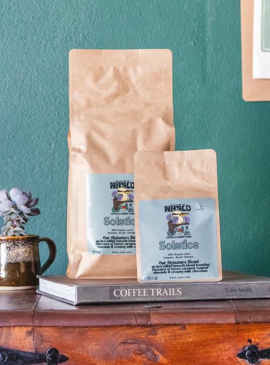 Roasted coffee beans from The Roast Collective. That's Whyld Coffee Roasters, Solstice Organic Blend. Image shows a 250g and a 1kg bag of Solstice blend next to each other, staged on a book titled "Coffee Trails", next to a succulent grown in a coffee cup. These are on the top of a wooden chest in front of a ocean green coloured background. The bags each have a duck egg blue coloured label centred on the lower half of the bag with the roaster's logo, blend name, and blend details on it. 