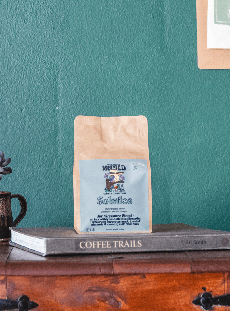 Roasted coffee beans from The Roast Collective. That's Whyld Coffee Roasters, Solstice Organic Blend. Image shows a brown 250g bag of Solstice blend staged on a book titled "Coffee Trails", next to a succulent grown in a coffee cup. These are on the top of a wooden chest in front of a ocean green coloured background. The bag has a duck egg blue coloured label centred on the lower half of the bag with the roaster's logo, blend name, and blend details on it. 