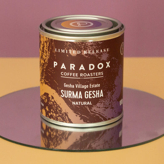 Roasted coffee beans. Paradox Coffee Roasters Limited Edition Single Origin: Gesha Village Estate, Surma Gesha. 100g tin on a mirror against a peach and mauve coloured background. Filter roast only. The Roast Collective