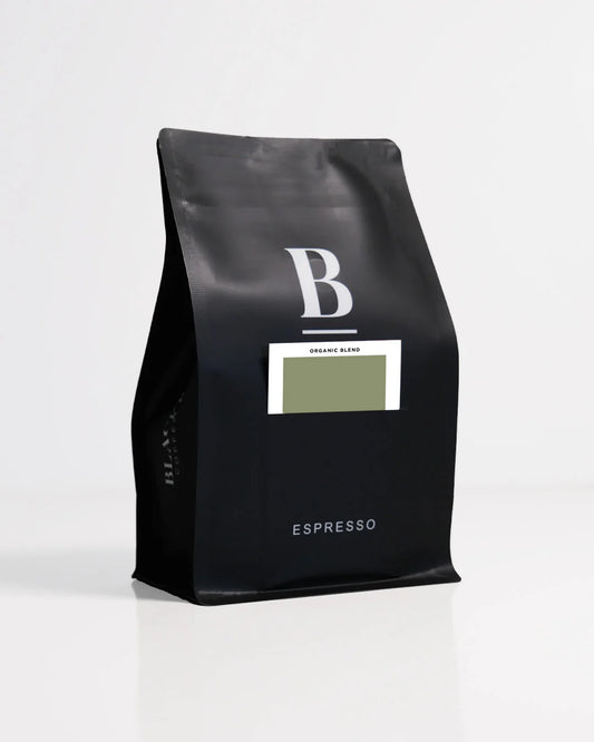 Roasted coffee beans. Blackboard Coffee Roasters, Blackboard Organic Espresso Blend. Image shows a black 250g bag against a white background. The bag features the Blackboard logo and a pouch on the front with a product information card poking out. The Roast Collective.