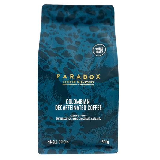 Roasted coffee beans from The Roast Collective. Paradox Coffee Roasters, Colombian Decaf Coffee. Image shows a 500g bag of the coffee against a white background. The bag is two-tone in colour, with navy blue and teal arranged in a reptile skin pattern. Paradox's logo is centred in the top half of the bag, with the blend name, tasting notes and weight underneath. 